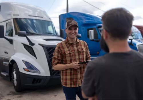A semi truck driver smiling and holding a digital tablet in his hands while talking to a mechanic
