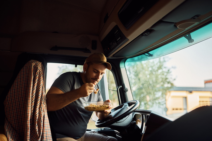 Male driver eating while making lunch break in truck's cabin. He's using Practical Tips for Healthier Eating on the Road